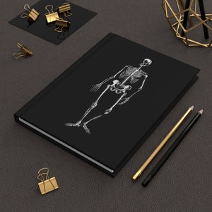 May include: A black journal with a white illustration of a human skeleton. The journal is rectangular and has a clean, minimalist design. Gold-colored binder clips and pencils are arranged around the journal on a dark gray surface.
