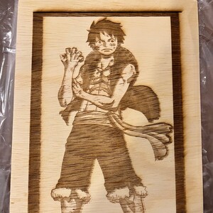 May include: A wood-burned image of a cartoon character with brown hair, wearing a red and white shirt and shorts, standing with one hand raised. The image is framed in a wood frame.