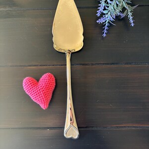 May include: A gold-colored cake server with a heart-shaped handle detail and a pink crocheted heart on a dark wood surface. A sprig of purple flowers is in the background. The server is approximately 10 inches long.
