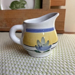May include: A small white ceramic creamer with a yellow, blue and green floral design. The creamer has a handle and a spout.