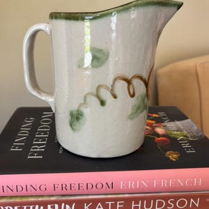 May include: A ceramic pitcher with a handle and spout, decorated with green and brown floral designs. The pitcher has a cream-colored base and a green rim. It sits on a stack of books with titles like "Finding Freedom" and "Pretty Fun".