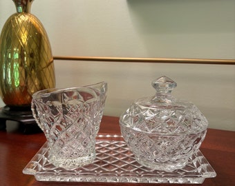 May include: A clear glass sugar bowl and creamer set with a diamond pattern on a clear glass tray. The set is on a wooden surface.
