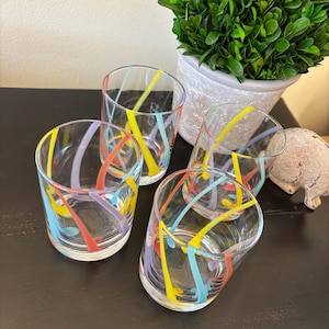 May include: Four clear drinking glasses with colorful, abstract stripes in yellow, red, blue, and purple. The glasses are arranged on a dark surface, with a potted plant in the background.