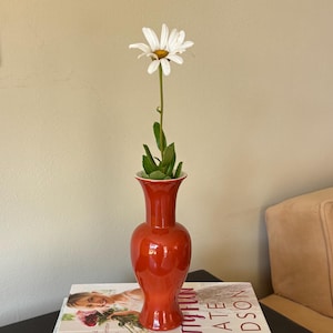 May include: A red vase holding a daisy with green leaves sits atop a stack of books. The books include "Pretty Fun Kate Hudson" and "Entertaining With Southern Living". The vase is a glossy, vibrant red.