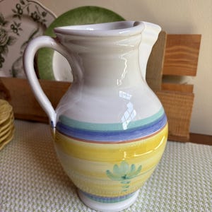 May include: A white ceramic jug with a curved handle and a wide spout. The jug is decorated with horizontal bands of yellow, blue, and green, and a painted floral design. The jug is likely used for serving beverages.