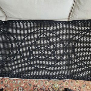 Puede incluir: Alfombra de crochet negra con un diseño de triple luna. La alfombra está hecha de un hilo fino y delicado y tiene una forma cuadrada.