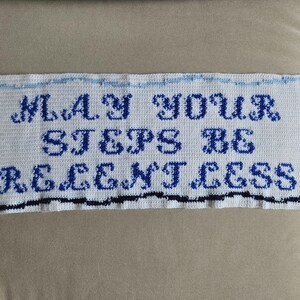 Peut inclure: Une pièce rectangulaire blanche, crochetée, avec les mots "MAY YOUR STEPS BE RELENTLESS" en bleu. La pièce a une bordure bleue et noire et est affichée sur une surface aux tons neutres.