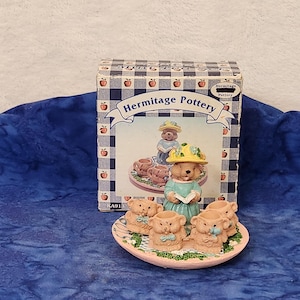 May include: A Hermitage Pottery figurine set featuring bear figures. The set includes a box with a picture of the figurine set, and a pink plate with several bear figurines. The box has a blue and white checkered pattern with apple illustrations.