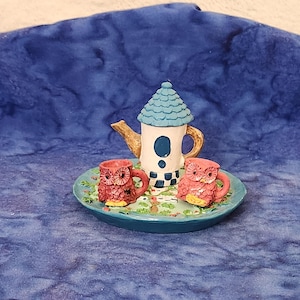 May include: A miniature ceramic tea set featuring a teapot and two owl-shaped cups on a small tray. The teapot has a blue roof and a white body with a black and white checkered pattern. The cups are red and pink.