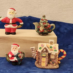 May include: A collection of Christmas-themed teapots and figurines. Includes a Santa Claus figurine, a Christmas tree teapot, and a gingerbread house teapot. The items are on a wooden box against a blue background.