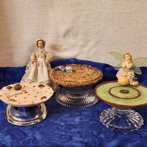 May include: A collection of decorative miniature display stands. One stand has a light-colored plate with brown spots, another has a woven top, and the third has a green and red design. A doll and a fairy figurine are also present.