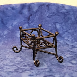 May include: A small, decorative black metal stand with a rectangular top and curved legs. The stand has a lattice design on the sides and is suitable for displaying small items. The stand is on a blue background.