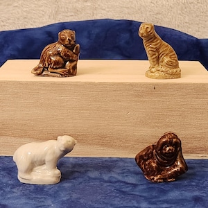 May include: Four small ceramic animal figurines: a brown monkey, a tan tiger, a white polar bear, and a brown gorilla. The figurines are displayed on a light brown wooden box against a dark blue background.