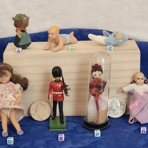 May include: A collection of vintage dolls and figurines, including a girl doll in a pink dress, a soldier figurine, a doll in a glass tube, a baby doll in a purple blanket, and a figurine of a person wearing a green hat and brown clothes.