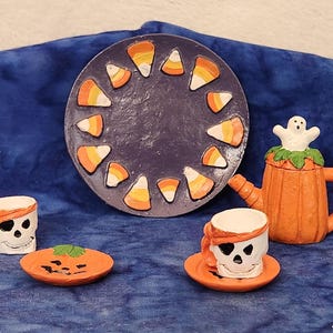 May include: A Halloween-themed ceramic set featuring a pumpkin teapot with a ghost on top, skull-shaped cups, and a candy corn decorated plate. The set includes a small pumpkin-shaped dish. The color palette includes orange, white, and purple.