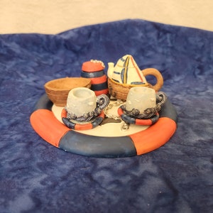 May include: A nautical themed tea set with a teapot shaped like a sailboat, two teacups, a creamer, and a sugar bowl. The set is on a red, white, and blue life preserver.