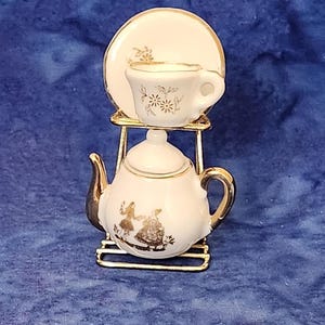 May include: A miniature white porcelain tea set with gold trim, including a teapot, teacup, and saucer, displayed on a gold-colored metal stand. The teapot features a decorative illustration of figures. The teacup and saucer have floral designs.