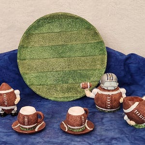 May include: A collection of football-themed ceramic items. Includes a large green plate resembling a football field, two coffee cups with saucers, a creamer, a sugar bowl, and a figurine of a football player.