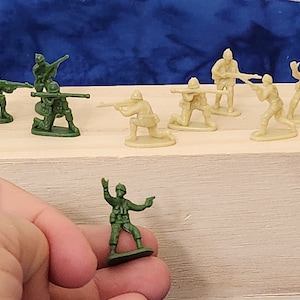 May include: A collection of green and tan plastic toy soldiers. The soldiers are posed in various action poses, including kneeling, standing, and aiming their rifles. The soldiers are arranged on a wooden surface.