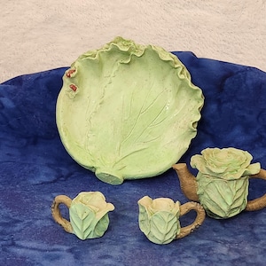 May include: A whimsical, light green ceramic tea set with a cabbage leaf-shaped serving plate, a teapot, and two teacups. The teapot and teacups are designed to resemble roses. The handles and spouts are brown. The set is on a blue surface.