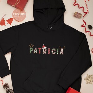 May include: Black hooded sweatshirt with a Christmas-themed design featuring the name "PATRICIA" in red, green, and white candy cane stripes. The letters are decorated with reindeer antlers and Santa hats.