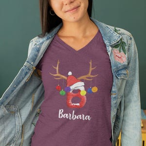 May include: Purple V-neck t-shirt with a Christmas reindeer design. The reindeer has a red Santa hat and is holding colorful ornaments. The text "Barbara" is printed below the reindeer.