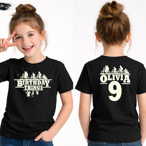 May include: Black t-shirt with a white graphic featuring bicycles, trees, and the text "BIRTHDAY THINGS" on the front. The back of the shirt displays the name "OLIVIA" and the number "9" in white.