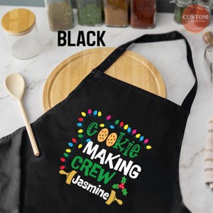May include: Black Christmas cookie making apron with a string of colorful lights, cookies, and the text "Cookie Making Crew Jasmine".