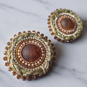 May include: Two decorative brooches with detailed designs. Each brooch has a central brown stone, encircled by layers of beads, crystals, and woven straw. The colour scheme includes browns, golds, and cream.