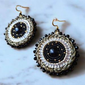 May include: A pair of large, round earrings with a black and white beaded design. The earrings have a gold-tone finish and feature a central black stone.