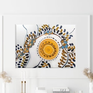 Puede incluir: Una pintura abstracta enmarcada con fondo blanco y un círculo amarillo central rodeado de formas marrones, azules y negras. La pintura está colgada en una pared blanca sobre un sofá blanco y una mesa blanca con un jarrón de flores secas.