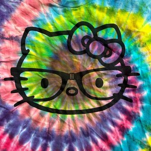 Puede incluir: Una camiseta tie-dye con un gr&aacute;fico negro de Hello Kitty con gafas. La camiseta tiene un patr&oacute;n de espiral arco&iacute;ris con tonos de rosa, morado, azul, verde y amarillo.