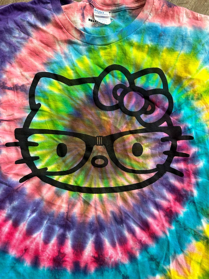 Puede incluir: Una camiseta tie-dye con un gr&aacute;fico negro de Hello Kitty con gafas. La camiseta tiene un patr&oacute;n de espiral arco&iacute;ris con tonos de rosa, morado, azul, verde y amarillo.