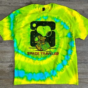 Può includere: Una maglietta tie-dye gialla, verde e blu con un'immagine di Snoopy in un costume da astronauta. Il testo "SPACE TRAVELER FIRST BEAGLE IN SPACE" è stampato sotto l'immagine.