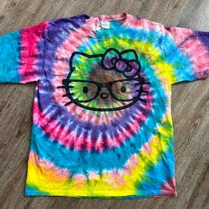 Puede incluir: Una camiseta tie-dye con un gr&aacute;fico de Hello Kitty en blanco y negro con gafas. La camiseta tiene un patr&oacute;n de espiral arco&iacute;ris en tonos de rosa, azul, amarillo y verde.