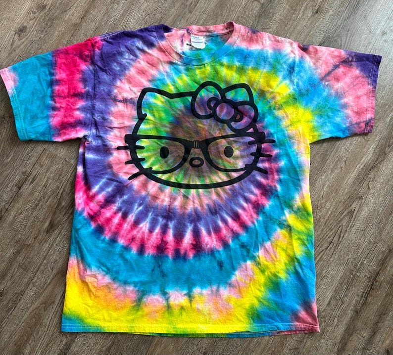 Puede incluir: Una camiseta tie-dye con un gr&aacute;fico de Hello Kitty en blanco y negro con gafas. La camiseta tiene un patr&oacute;n de espiral arco&iacute;ris en tonos de rosa, azul, amarillo y verde.