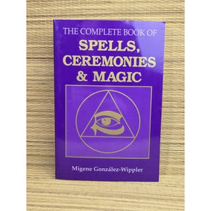 May include: A purple book titled The Complete Book of Spells, Ceremonies & Magic by Migene González-Wippler. The cover features gold text and a gold graphic of an eye inside a triangle and circle. The book has a glossy finish and rectangular shape.
