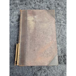 May include: An antique book with a worn, brown leather cover. The spine and corners are a darker brown, and the pages show signs of age. The book's cover has a faded appearance, suggesting it has been well-used over time.