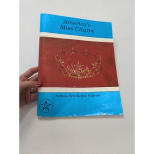 May include: A vintage book titled "America's Miss Charm" with a blue cover and a red background. The cover features a gold crown illustration. The text "National Scholarship Pageant" is also visible.