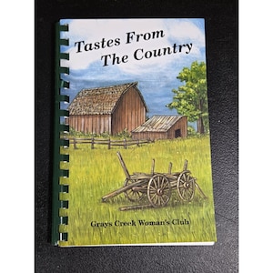 May include: A spiral-bound cookbook titled Tastes From The Country by Grays Creek Woman's Club. The cover features a painting of a wooden barn, a smaller shed, and a wooden wagon in a green field under a blue sky with white clouds.