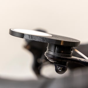 Op de afbeelding: Een zwarte en zilveren fietsbevestiging met een ronde, getextureerde basis en een zwenkarm. De bevestiging is ontworpen om een camera of ander apparaat vast te houden.