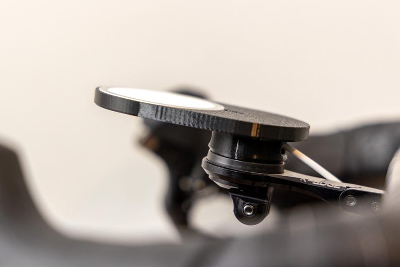 Op de afbeelding: Een zwarte en zilveren fietsbevestiging met een ronde, getextureerde basis en een zwenkarm. De bevestiging is ontworpen om een camera of ander apparaat vast te houden.