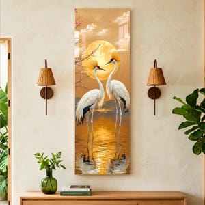May include: A vertical rectangular wall art print featuring two white cranes with red crowns standing in shallow golden water. A large, bright yellow full moon hangs in the sky behind them, with thin tree branches and soft clouds visible.