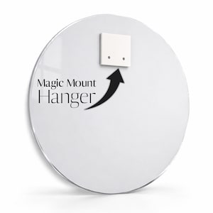 May include: A circular, clear acrylic panel featuring a white square mounting bracket attached to the upper centre. The text Magic Mount Hanger is printed in black, accompanied by a black arrow pointing towards the mounting bracket.