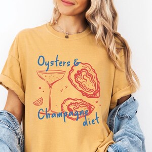 May include: A yellow t-shirt with the text "Oysters & Champagne diet" in blue and red lettering. The shirt features a graphic of a champagne glass, a lemon slice, and two oysters.
