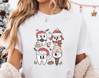 May include: White t-shirt featuring a whimsical Christmas-themed design. The graphic includes four cartoon teeth characters wearing Santa hats and scarves, with festive elements like cookies and candy canes. The model is wearing red and white plaid pajama pants.
