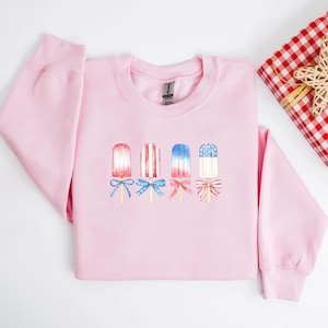 May include: A light pink sweatshirt featuring a watercolor design of four popsicles in red, white, and blue. Each popsicle has a bow at the bottom. The sweatshirt is a crew neck style.