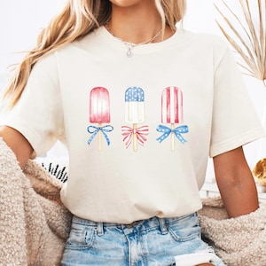May include: Cream-colored t-shirt featuring three watercolor popsicle designs. Each popsicle has red, white, and blue accents, with bows at the base. The shirt is paired with blue denim jeans.