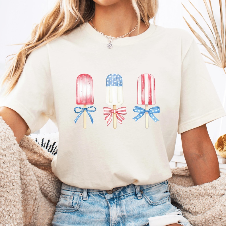 May include: Cream-colored t-shirt featuring three watercolor popsicle designs. Each popsicle has red, white, and blue accents, with bows at the base. The shirt is paired with blue denim jeans.
