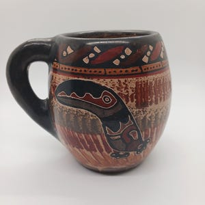 May include: A ceramic mug with a black handle and rim, featuring a brown and tan body with a tribal design. The mug has a dark brown and red pattern around the top and a stylized bird or animal figure on the front.